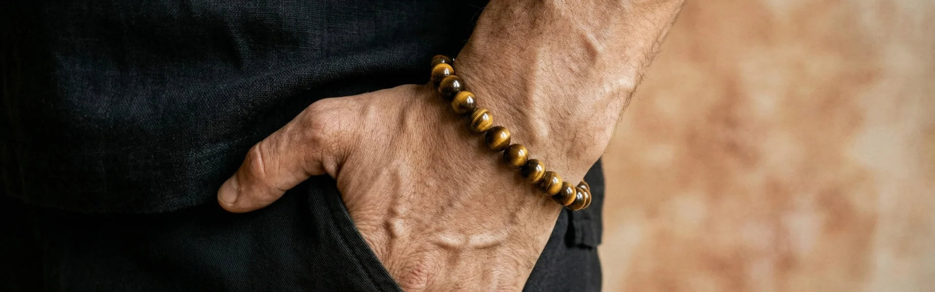 Gros plan sur un bracelet homme en Œil de Tigre. Perles rondes en pierres naturelles chatoyantes au poignet, sur fond texturé ocre. Un accessoire artisanal authentique.
