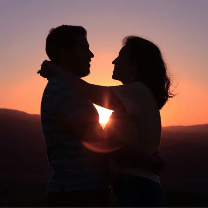Une photo en silhouette d'un couple s'embrassant tendrement devant un coucher de soleil chaleureux et doré. Le soleil bas se trouve entre eux, créant un flare dramatique et une lumière dorée. Ils se regardent. La scène évoque la romance, la proximité et la connexion profonde, même à distance, entre les partenaires. Le coucher de soleil dramatique symbolise la fin de la distance et le début de l'union.