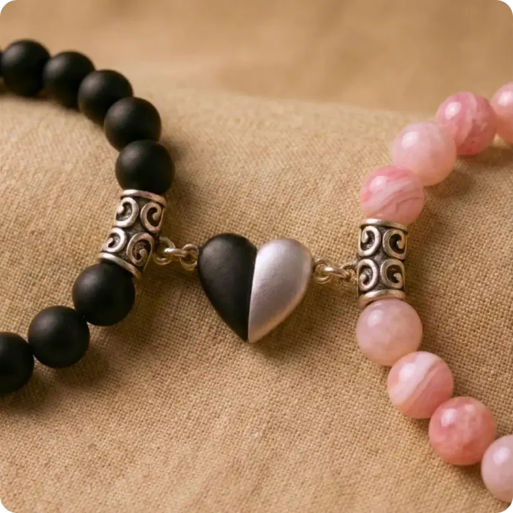 Détail d'un duo de bracelets couple distance symbolisant l'amour. Un bracelet en perles d'Onyx noir mat est relié à un bracelet en Jade rose véritable par un pendentif cœur aimanté, posés sur un tissu texturé beige.