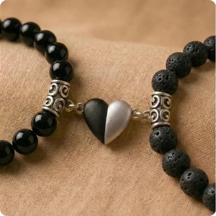 Gros plan sur un duo de bracelets couple distance en pierres naturelles. À gauche, des perles en Onyx noir poli, à droite, de la Pierre de Lave brute poreuse. Les deux bracelets sont connectés au centre par un pendentif demi-cœur aimanté bicolore noir et argent.
