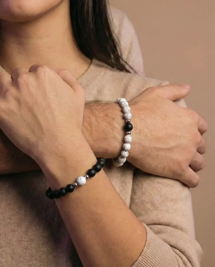 Vue rapprochée de deux mains de couple portant des bracelets assortis. Un bracelet est composé de perles d'onyx noir mat avec une perle centrale en howlite blanche et des entretoises argentées. L'autre bracelet est composé de perles d'howlite blanche avec une perle centrale en onyx noir mat et des entretoises argentées. Les perles sont des pierres naturelles de 8mm. Les mains se touchent, symbolisant la connexion et l'union, même à distance.